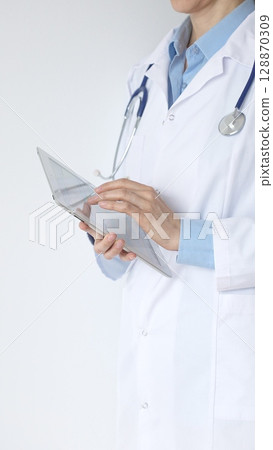 Unknown female doctor, wearing white coat over a blue blouse and a stethoscope, is using a digital tablet, suggesting innovative healthcare practices in medicine. Health care and medicine concept 128870309
