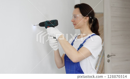 Woman worker wearing blue overall, safety glasses and gloves, is using cordless drill to make hole in white wall during renovation work. Horizontal portrait view 128870314