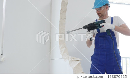 Man construction worker wearing blue overalls, hard hat and protective white gloves, is demolishing white wall with rotary hammer drill, generating dust, close up horizontal view. Renovation concept Man construction worker wearing blue overalls, hard hat and protective white gloves, is demolishing white wall with rotary hammer drill, generating dust, close up horizontal view. Renovation concept 128870315