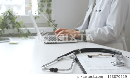 Stethoscope and medical records are on the desk opposite the doctor is typing on a laptop with a in a medical office, suggesting online healthcare or telemedicine services. Medicine concept 128870319