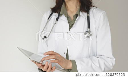 Unknown professional woman doctor, wearing white coat over a green blouse and a stethoscope, is using a digital tablet, suggesting innovative healthcare practices in medicine. Health care concept Unknown professional woman doctor, wearing white coat over a green blouse and a stethoscope, is using a digital tablet, suggesting innovative healthcare practices in medicine. Health care concept 128870323