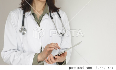 Unknown professional woman doctor, wearing white coat over a green blouse and a stethoscope, is using a digital tablet, suggesting innovative healthcare practices in medicine. Health care concept Unknown professional woman doctor, wearing white coat over a green blouse and a stethoscope, is using a digital tablet, suggesting innovative healthcare practices in medicine. Health care concept 128870324
