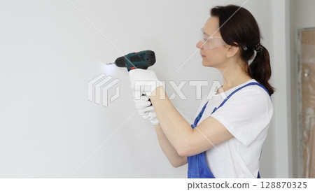 Woman worker wearing blue overall, safety glasses and gloves, is using cordless drill to make hole in white wall during renovation work. Horizontal portrait view 128870325