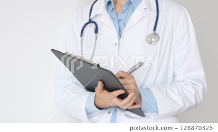 Unknown female doctor wearing stethoscope and white coat over blue blouse is standing and writing on a black clipboard, representing medical expertise and patient care. Medicine concept 128870326