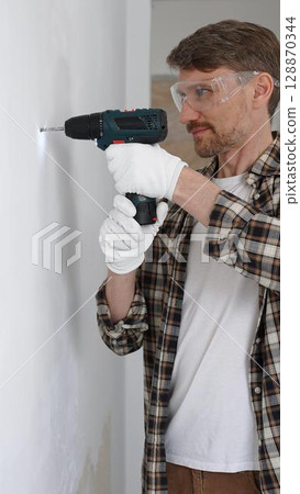 Middle aged male worker wearing checked shirt and white gloves, is drilling hole in a wall with cordless drill during home renovation work. Vertical portrait view 128870344