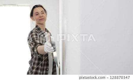 Female construction professional wearing checkered shirt is smoothing white wall surface, applying putty with professional spatula technique, showcasing skilled interior renovation work 128870347