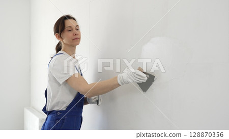Construction worker woman spreading finishing putty on white wall, wearing protective gloves while smoothing surface with putty knife for precise home repair Construction worker woman spreading finishing putty on white wall, wearing protective gloves while smoothing surface with putty knife for precise home repair 128870356