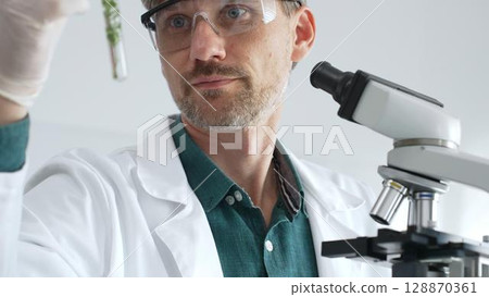 Adult male scientist in mask and gloves holding test tube with green plant sample, conducting laboratory research with a microscope. Medicine, health care and science concept 128870361