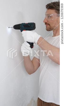 Middle aged male worker wearing white t-shirt and protective gloves, is drilling hole in a wall with cordless drill during home renovation work. Portrait view 128870369