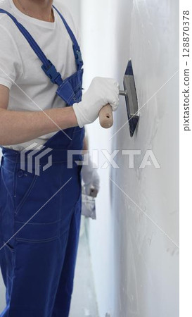 Professional construction worker wearing blue work overall is using a finishing trowel to carefully apply plaster on a wall, demonstrating expertise in home renovation Professional construction worker wearing blue work overall is using a finishing trowel to carefully apply plaster on a wall, demonstrating expertise in home renovation 128870378