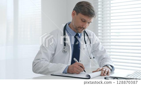 A male doctor in a lab coat with a stethoscope sits at a table in a medical office and uses a laptop while taking notes on a clipboard for patient care and treatment A male doctor in a lab coat with a stethoscope sits at a table in a medical office and uses a laptop while taking notes on a clipboard for patient care and treatment 128870387