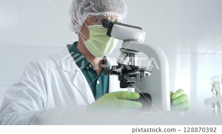 Medical scientist wearing protective mask, cap and green gloves using microscope doing scientific research in laboratory, developing vaccine or medication. Medicine, healthcare and science concept 128870392
