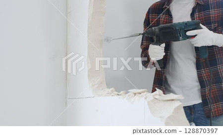 Close up of unknown male construction worker wearing red checkered shirt jeans and protective gloves, is demolishing white wall with rotary hammer drill, generating dust. Renovation concept Close up of unknown male construction worker wearing red checkered shirt jeans and protective gloves, is demolishing white wall with rotary hammer drill, generating dust. Renovation concept 128870397