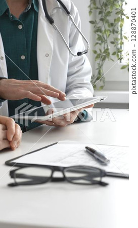 Doctor man in lab coat over a green shirt, and stethoscope is using tablet while consulting patient in modern medical office. Medicine and health care concept Doctor man in lab coat over a green shirt, and stethoscope is using tablet while consulting patient in modern medical office. Medicine and health care concept 128870401