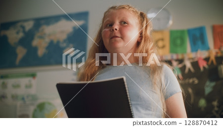 Primary School Girl with Notebook Speaking, Showcasing Knowledge of Geography in Front of Classroom 128870412