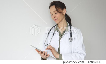 Doctor woman wearing white medical coat over green blouse, stethoscope, is reviewing patient records on digital tablet, standing in medical workspace with natural lighting. Medicine concept 128870413