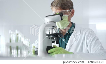 Adult male scientific professional analyzing sample through microscope, wearing green protective gear in sterile laboratory setting. Medicine, healthcare and science concept Adult male scientific professional analyzing sample through microscope, wearing green protective gear in sterile laboratory setting. Medicine, healthcare and science concept 128870430