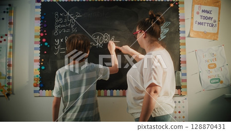 Smart Boy Solving Mathematical Problems on the Chalkboard. Female Teacher Checking Answers Smart Boy Solving Mathematical Problems on the Chalkboard. Female Teacher Checking Answers 128870431