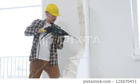 Man construction worker wearing beige checkered shirt , yellow hard hat and protective gloves, is demolishing white wall with rotary hammer drill, generating dust, medium long shot. Renovation concept 128870451