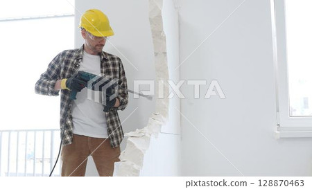 Man construction worker wearing beige checkered shirt , yellow hard hat and protective gloves, is demolishing white wall with rotary hammer drill, generating dust, medium long shot. Renovation concept Man construction worker wearing beige checkered shirt , yellow hard hat and protective gloves, is demolishing white wall with rotary hammer drill, generating dust, medium long shot. Renovation concept 128870463