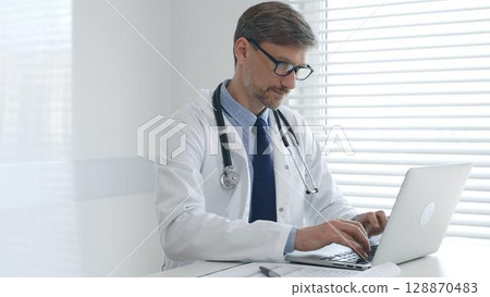 Focused mature male doctor wearing eyeglasses and lab coat with stethoscope is typing on laptop keyboard while sitting at desk in bright medical office 128870483
