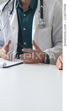 Doctor man wearing medical coat over green shirt, and stethoscope is gesturing with hands while explaining diagnosis to patient during medical consultation in clinic office. Medicine and health care Doctor man wearing medical coat over green shirt, and stethoscope is gesturing with hands while explaining diagnosis to patient during medical consultation in clinic office. Medicine and health care 128870486