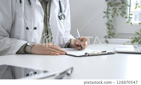Professional female doctor wearing white lab coat and stethoscope documenting patient information on clipboard near laptop in clinic workspace. Medicine and health care concept 128870547