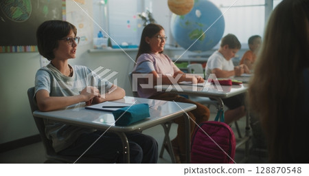 Elementary School Students Sitting at the Desks, Raising Hands to Provide Correct Answer 128870548