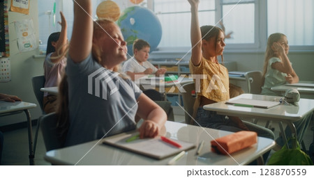 Primary School Children Sitting at the Desks, Listening to Lecture, Raising Hands to Give Correct Answer Primary School Children Sitting at the Desks, Listening to Lecture, Raising Hands to Give Correct Answer 128870559