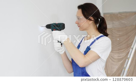 Professional female worker wearing blue overall, safety glasses and gloves, is using cordless drill to make hole in white wall during renovation work. Portrait view 128870603