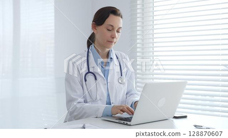 Female doctor working on laptop in white lab coat and stethoscope, sitting at desk in bright, sunlit medical office. Medicine and healthcare concept Female doctor working on laptop in white lab coat and stethoscope, sitting at desk in bright, sunlit medical office. Medicine and healthcare concept 128870607