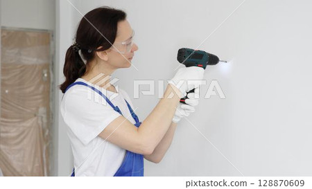 Woman worker wearing blue overall, safety glasses and gloves, is using cordless drill to make hole in white wall during renovation work. Horizontal portrait view 128870609