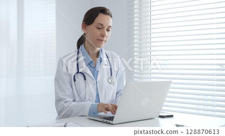 Professional female physician working on laptop, wearing white lab coat and stethoscope, seated at desk in bright, sunlit medical workspace. Medicine and health care concept Professional female physician working on laptop, wearing white lab coat and stethoscope, seated at desk in bright, sunlit medical workspace. Medicine and health care concept 128870613
