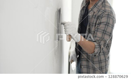 Unknown male construction worker wearing gloves and plaid shirt is using finishing trowel to plaster a white wall, applying a layer of plaster to create a smooth surface during renovation project 128870629