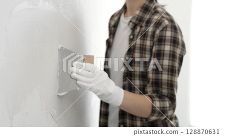 Unknown female construction worker wearing protective gloves and checkered shirt is spreading plaster on white wall during renovation project, smoothing surface with putty knife 128870631
