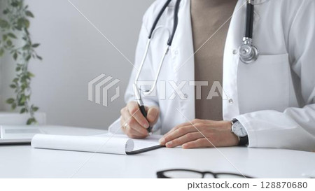 Male doctor working at desk, writing prescription, wearing white lab coat and stethoscope in clinical workspace, close up. Medicine and health care concept Male doctor working at desk, writing prescription, wearing white lab coat and stethoscope in clinical workspace, close up. Medicine and health care concept 128870680