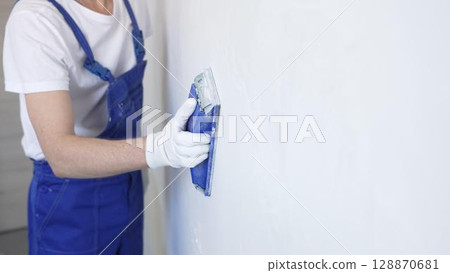 Unknown male construction worker wearing protective white gloves and blue construction coveralls, is sanding white wall with blue hand block, preparing smooth surface during interior home renovation Unknown male construction worker wearing protective white gloves and blue construction coveralls, is sanding white wall with blue hand block, preparing smooth surface during interior home renovation 128870681