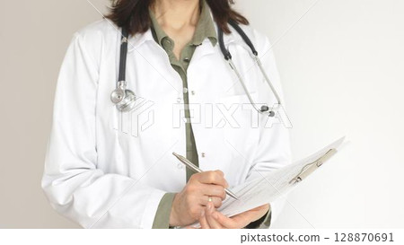 Female doctor, wearing a stethoscope and a medical coat over a green blouse, is standing and writing on a white clipboard, representing medical expertise and patient care. Medicine concept. 128870691