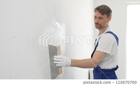 Male construction worker wearing protective gloves and blue construction coveralls, spreading plaster smoothly across wall using professional drywall taping knife during renovation project. Portrait 128870700