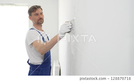Middle aged male construction worker wearing gloves and blue overalls is using a putty knife to apply plaster on a white wall, performing home renovation Middle aged male construction worker wearing gloves and blue overalls is using a putty knife to apply plaster on a white wall, performing home renovation 128870718
