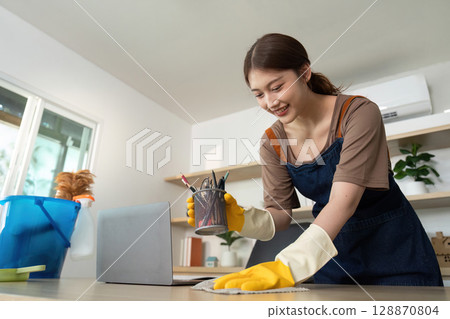 Home Cleaning. Young woman using spray cleaner and cloth to tidy up a modern workspace. Home Cleaning. Young woman using spray cleaner and cloth to tidy up a modern workspace. 128870804