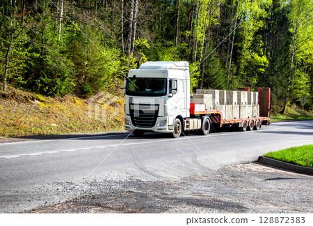Modern truck with a lowboy semitrailer transports concrete blocks along a country road against the backdrop of a forest in summer. Beautiful nature, industry. Copy space for text 128872383