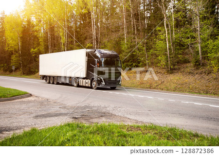 A truck with a refrigerated semi-trailer transports refrigerated cargo along a country road against the backdrop of beautiful nature and forest in summer. Copy space for text A truck with a refrigerated semi-trailer transports refrigerated cargo along a country road against the backdrop of beautiful nature and forest in summer. Copy space for text 128872386