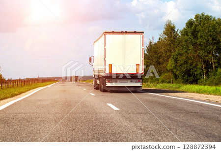 A truck with a semi-trailer transports a load of construction materials along the highway in the summer against the backdrop of the sun. Concept of working as a truck driver, copy space for text 128872394