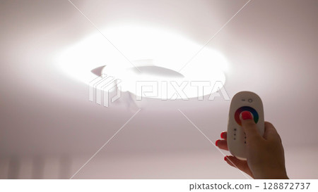 Remote control in hand against the background of a ceiling diode chandelier. Concept of remote control of chandelier lighting Remote control in hand against the background of a ceiling diode chandelier. Concept of remote control of chandelier lighting 128872737