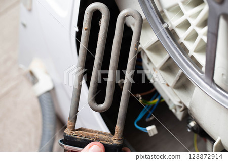 An electric heating element with scale and deposits against the background of a washing machine. Concept of repair and replacement of spare parts, close-up 128872914