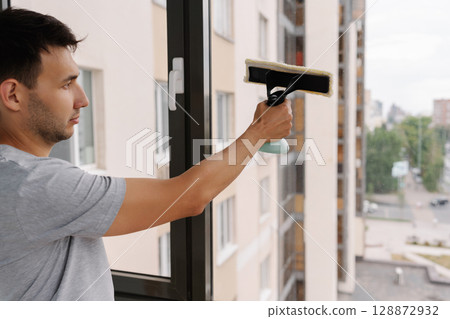 Janitor male spraying cleaning solution onto large window and wiping clean with squeegee, ensuring spotless and streak-free finish in modern building. Concept of cleanliness and housekeeping. Janitor male spraying cleaning solution onto large window and wiping clean with squeegee, ensuring spotless and streak-free finish in modern building. Concept of cleanliness and housekeeping. 128872932