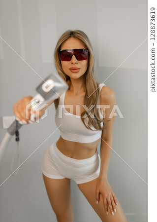 Woman tunes a laser hair removal machine. She holds a working part of the epilator in her hands and poses for a photo. Body Care. Underarm Laser Hair Removal. Woman tunes a laser hair removal machine. She holds a working part of the epilator in her hands and poses for a photo. Body Care. Underarm Laser Hair Removal. 128873619