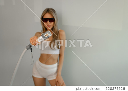 Woman tunes a laser hair removal machine. She holds a working part of the epilator in her hands and poses for a photo. Body Care. Underarm Laser Hair Removal. 128873626
