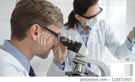 A researcher wearing glasses examines a sample under a microscope, a lab technician wearing a protective mask stands nearby, both in white lab coats during the study. Medicine and Science A researcher wearing glasses examines a sample under a microscope, a lab technician wearing a protective mask stands nearby, both in white lab coats during the study. Medicine and Science 128873803